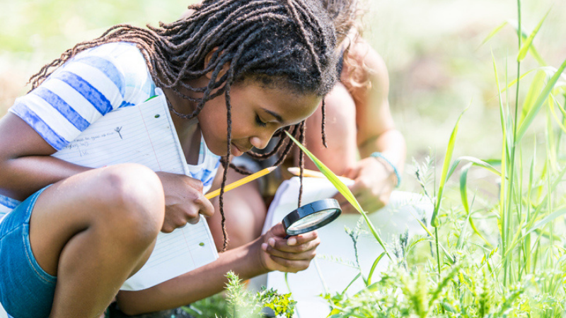 Chica afroamericana haciendo actividad de insectos al aire libre, usando una lupa para agacharse y explorar en la hierba
