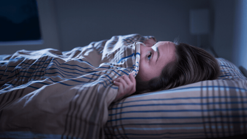 Una mujer joven yace en la cama con una computadora a rayas azules y marrones que cubre la mitad de su rostro.