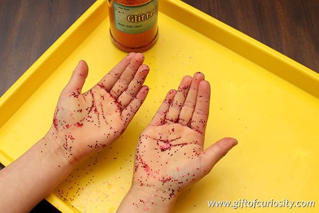 Las manos de un joven estudiante cubiertas con purpurina roja en una bandeja amarilla